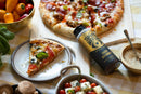 A Spice Witch Hot Honey Squeeze Bottle sits next to a pizza slice on a plate, a whole pizza on a wooden board, and bowls of cherry tomatoes and colorful peppers atop a checkered tablecloth.