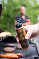 A hand holds a Spice Witch Hot Honey Squeeze Bottle over a grill of sizzling sausages and burgers, while someone with a red cup stands blurred in the background.