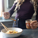 In a kitchen, someone holds a jar of Spice Witch Chili Crisp Original Flavor, packed with crispy garlic, and drizzles it from a spoon onto noodles with chopsticks.