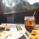 A jar of Spice Witch Chili Crisp Original Flavor by Spice Witch, with crispy garlic and a spoon inside, sits on a table next to a plate of partially eaten bread and a fork, sunlight streaming through the window in the background.