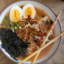 A bowl of ramen soup topped with sliced boiled eggs, green onions, dried seaweed, crispy garlic, meat slices, and a spoonful of Spice Witch Chili Crisp Original Flavor by Spice Witch. Chopsticks rest on the bowl’s edge.