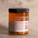 A small jar of Spice Witch Chili Crisp Original Flavor, with a black lid and clear label displaying ingredients, suggested uses, and nutrition facts in white text. The jar from Spice Witch rests on a neutral surface.