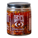 A glass jar of Spice Witch Chili Crisp Original Flavor by Spice Witch, with a mystical eye logo. The label says "100% magical, 0% boring." Inside, red chili flakes and crispy garlic in avocado oil are clearly visible.