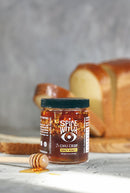 A jar of Spice Witch Chili Crisp Spicy Honey Flavor by Spice Witch sits beside sliced bread, with a honey dipper of spicy honey in front. The background is softly blurred, highlighting the crunchy condiment.