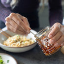 A person holds a jar of Spice Witch Chili Crisp Spicy Honey Flavor by Spice Witch and scoops some onto noodles garnished with green onions, using chopsticks.