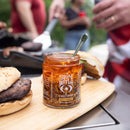 An open jar of Spice Witch Chili Crisp Spicy Honey Flavor from Spice Witch, with a spoon inside, sits on a wooden board beside a burger bun and patty while people grill in the background.