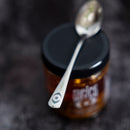 A silver spoon with a small eye illustration on its handle rests atop a closed jar of Spice Witch Chili Crisp Spicy Honey Flavor by Spice Witch, featuring a black lid and set against a dark, blurred background.