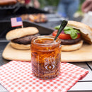 A jar of Spice Witch Chili Crisp Spicy Honey Flavor by Spice Witch sits with a spoon on a red checkered napkin, ready to be drizzled on burgers. An American flag toothpick decorates the background, hinting at its bold, crunchy flavor.