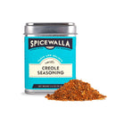 A blue Spicewalla Creole Seasoning tin with black and white accents and a metal lid sits next to a small pile of reddish-brown spice blend, perfect for classic Louisiana dishes like gumbo.