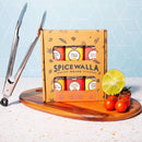 Spicewalla grill and roasting collection spice set on a wooden board with kitchen tools and ingredients against a light blue tiled wall.