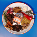 A white plate features the Spicewalla Jerk Seasoning tin, surrounded by grilling spices like shallots, garlic, thyme, rosemary, red chili, brown sugar, coriander seeds, cinnamon sticks, nutmeg, black peppercorns, and allspice.