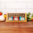 The Spicewalla Kitchen Essentials 3-Pack—Sicilian Sea Salt, Crushed Red Pepper, and Black Pepper—rests on a wooden counter with garlic, herbs, and veggies against a white tiled backdrop.