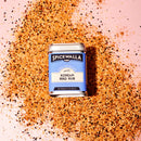 A tin of Spicewalla Korean BBQ Rub rests on a layer of reddish-brown sweet and savory seasoning, its blue and white label standing out against a pink background, hinting at bold flavors for grilling.