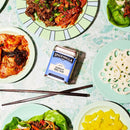 A tin of Spicewalla Korean BBQ Rub by Spicewalla is displayed with plates of vibrant Korean dishes and grilled meats, including kimchi, lotus root, pickled radish, and lettuce wraps, offering a sweet-savory seasoning experience. Chopsticks rest nearby.