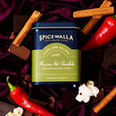 A tin of Spicewalla Mexican Deluxe Hot Chocolate is surrounded by cinnamon sticks, red chili peppers, chocolate pieces, and marshmallows on a dark purple velvet background.