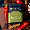 A tin of Spicewalla Mexican Deluxe Hot Chocolate rests atop chocolate bars, surrounded by red chili peppers, cinnamon sticks, and chunks of rich chocolate.