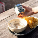 A hand holds a can of Spicewalla Mignonette Blend for Seafood above a bowl of dip with a spoon, next to potato chips on a tray atop a wooden table.
