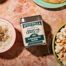 A tin of Spicewalla Mignonette Blend for Seafood by Spicewalla rests on a marble surface with popcorn, dip, and bread—perfect as an oyster mignonette seasoning or a flavorful topping for seafood dishes.