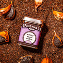 A jar of Spicewalla Minced Black Garlic by Spicewalla rests on a layer of black garlic granules, with peeled black garlic cloves nearby, showcasing this rich umami seasoning.