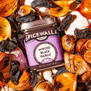 A jar of Spicewalla Minced Black Garlic by Spicewalla sits amid scattered garlic cloves and black garlic pieces, brightly lit in warm tones to highlight this unique umami seasoning.