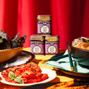Three tins of Spicewalla Minced Black Garlic are stacked among bowls of mussels, noodles, and a plate of roasted tomatoes and basil. The Spicewalla seasoning adds rich umami depth to the dishes on a wooden table with a red curtain backdrop.