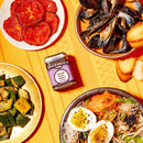A vibrant spread on a yellow tablecloth showcases the rich taste of Spicewalla Minced Black Garlic, paired with roasted tomatoes, mussels and toasted bread, cucumber salad, and a noodle bowl topped with eggs and fresh herbs.