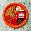 An orange plate on a green marbled surface displays two red chili peppers, black peppercorns, fresh thyme, ground spices, garlic cloves, and a tin of Spicewalla N'Asheville Hot Chicken Seasoning.