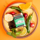 A round plate with Spicewalla Pescado Verde Seafood Seasoning by Spicewalla, surrounded by green chilies, red shallots, black peppercorns, garlic, cilantro, lime wedge, and orange slice highlights this vibrant citrus herb seafood blend.