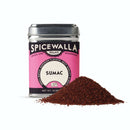 A tin of Spicewalla Sumac sits behind a small pile of reddish-brown ground sumac, prized for its tart citrus flavor in Middle Eastern cooking, on a white background.