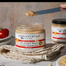 A jar of Terrapin Ridge Farms Hot Pepper Bacon Cream Cheese Dip rests on a beige towel with a knife spreading the dip. Another jar and fresh tomatoes appear in the background, all on a white surface. Brand: Terrapin Ridge Farms.