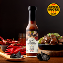 A bottle of Texan Heartland Black Garlic Arbol Hot Sauce stands on a wooden surface, surrounded by dried and fresh chili peppers, black garlic, and a dish of food. A 2026 European Hot Sauce Awards silver medal is displayed.