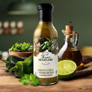 A bottle of Texan Heartland Green Chile Chimichurri Marinade & Sauce sits on a wooden table with fresh parsley, green chile, half a lime, and olive oil nearby—showcasing bold Southwestern flavors in a blurred kitchen setting.