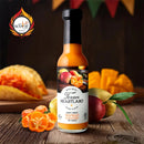 A bottle of Texan Heartland Honey Ginger Mango Habanero Hot Sauce rests on wood, surrounded by mango slices, habanero peppers, and a taco. The Scovie Award logo in the corner spotlights this spicy-sweet Texan Heartland condiment.