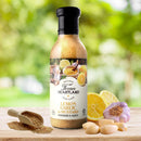 A bottle of Texan Heartland Lemon, Garlic & Mustard Marinade & Sauce rests on a wooden surface, surrounded by garlic cloves, a garlic bulb, lemon halves, and mustard seeds, with a blurred green outdoor background.