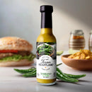 A bottle of Texan Heartland Serrano Sunfire Hot Sauce sits on a table with fresh serrano peppers, a burger, and chips. Its label features green peppers and a rustic design, reflecting the bold Texas flavor of Texan Heartland.