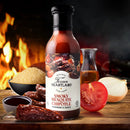 A bottle of Texan Heartland Smoky Mesquite Chipotle Marinade & Sauce sits among chipotle peppers, brown sugar, a tomato, onion, oil, and a bowl of sauce with fire blazing behind—capturing the bold essence of Texas BBQ.