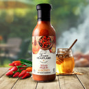 A bottle of Texan Heartland Texas Pequin & Honey Mop Sauce & Marinade rests on a wooden table beside fiery pequin peppers and honey, ideal for adding a spicy-sweet kick to your marinades, with a softly blurred outdoor backdrop.