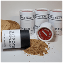 Four Texas Salt Co. Chipotle Cocktail Beer Salt containers are shown, one tipped over and spilling smoky salt onto a white surface—ideal for rimming margaritas or craft cocktails. The Texas Salt Co logo is visible on each lid.