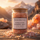 A Texas Salt Co. Himalayan Pink Extra Coarse Salt glass jar sits on wood, surrounded by pink salt crystals, with mountains and a bright sky in the background.
