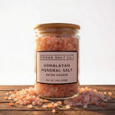 A glass jar labeled Texas Salt Co Himalayan Pink Extra Coarse Salt sits on a wooden surface, filled with extra coarse pink salt crystals and surrounded by scattered Himalayan pink salt pieces.