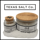 Two glass jars of Texas Salt Co Oz Australian Salt—one with a Texas-shaped cork lid and one with a bamboo lid—rest on coarse sea salt, with the Texas Salt Co logo displayed above them.