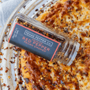 A bottle of Texas Salt Co. Pecan Smoked Crushed Red Pepper is tipped over a cheesy pizza with smoked chili peppers, served on a round white plate.