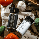 The Texas Salt Co. Salt & Pepper Set, an essential seasoning duo for everyday use, rests on a cloth amid fresh vegetables and garlic in natural light.