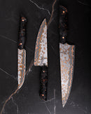 The Folded Steel Hellebore 3-Piece CuMai Knife Set with AUS10 Damascus steel is displayed on black marble, featuring dark marbled handles and intricately patterned silver and copper blades.