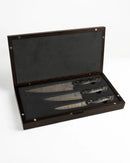 A dark wooden case with a foam-lined interior holds Folded Steel’s Hellebore 3-Piece CuMai Knife Set with AUS10 Damascus Steel, each knife featuring a marbled pattern and unique handles, displayed diagonally on a white background.