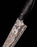 A close-up of Folded Steel’s Hellebore 3-Piece CuMai Knife Set showcases an AUS10 Damascus blade with swirling silver, black, and copper patterns and a dark handle accented by copper flecks—all set against a black background.