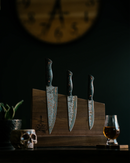 Three Folded Steel Hellebore 3-Piece CuMai AUS10 Damascus Steel knives with dark handles sit on a wooden block, near a glass of whiskey, skull-shaped container, and plant. A large blurred clock hangs in the background.