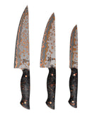 The Folded Steel Hellebore 3-Piece CuMai Knife Set features AUS10 Damascus steel blades with intricate patterns and black handles with copper accents, displayed upright side by side against a white background.
