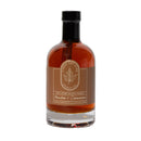A clear glass bottle with a black cap contains Under The Bark Syrup Co Bourbon + Cinnamon Maple Syrup. The brown and cream label features a maple leaf design and states "100% Pure Maple Syrup" by Under The Bark Syrup Co.