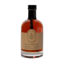 A clear glass bottle of Under The Bark Syrup Co Bourbon + Vanilla Maple Syrup, 12.5 fl oz, features a beige label and black cap. Made in Minnesota—makes an ideal gourmet gift for syrup lovers.
